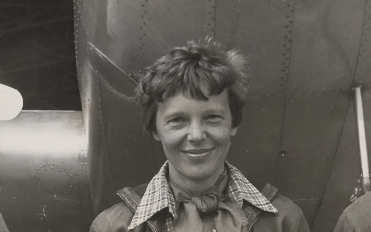 Black-and-white archive photo of Amelia Earhart smiling as she stands under the nose of her Lockheed Electra 10-E
