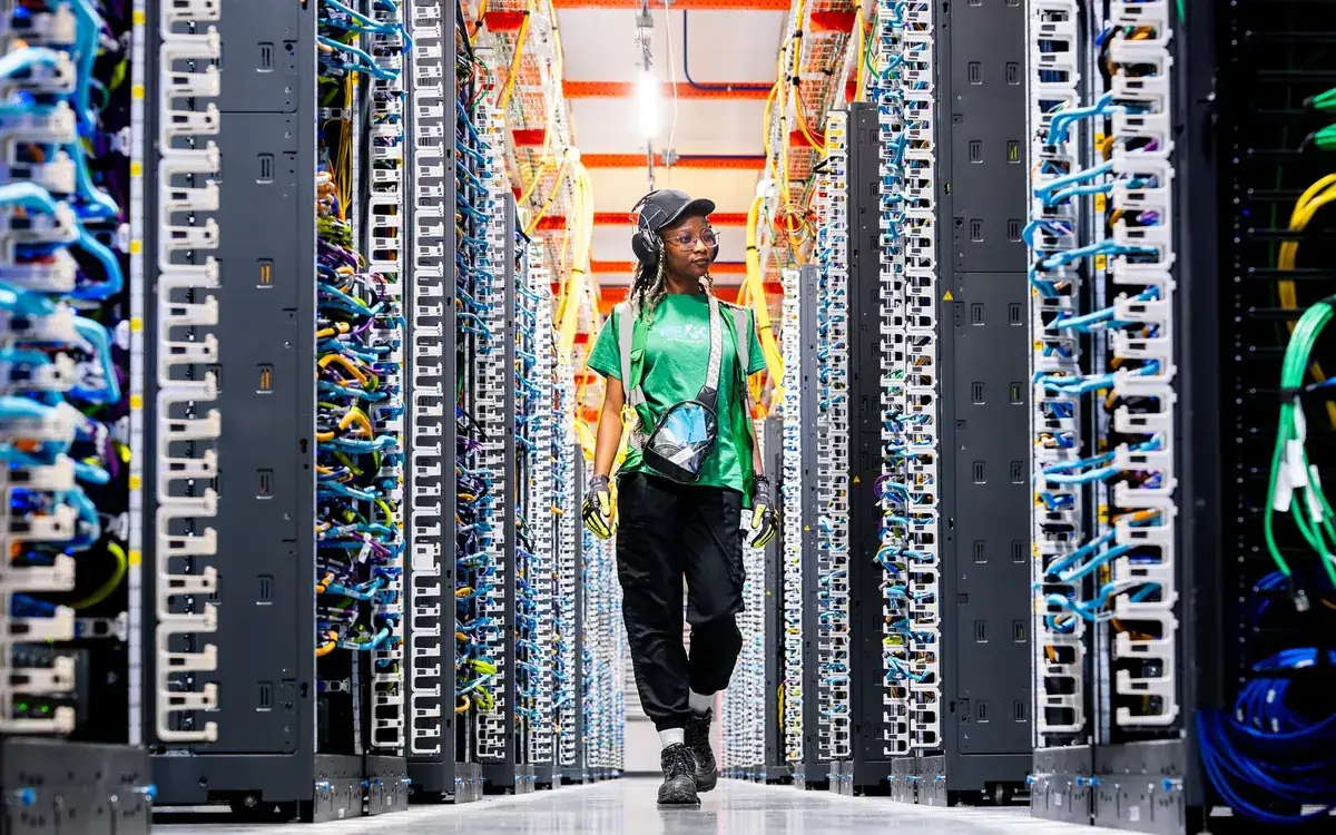 An Amazon data centre aisle lined with server racks and blue and green network cables, with a technician walking between them
