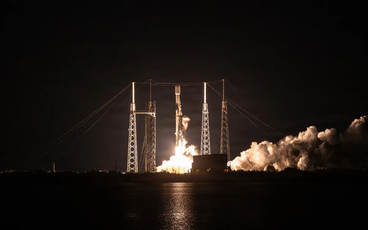 A Falcon 9 rocket lifts off at night from Cape Canaveral, its exhaust lighting the launch tower and steam cloud (earlier Bandwagon-4 mission, illustrative)