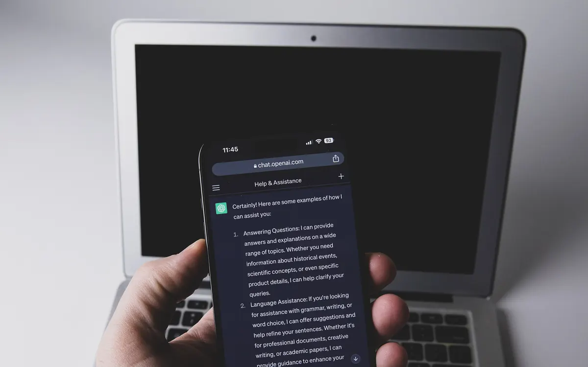 A hand holding a smartphone with the ChatGPT app open in front of a laptop (illustrative photo)