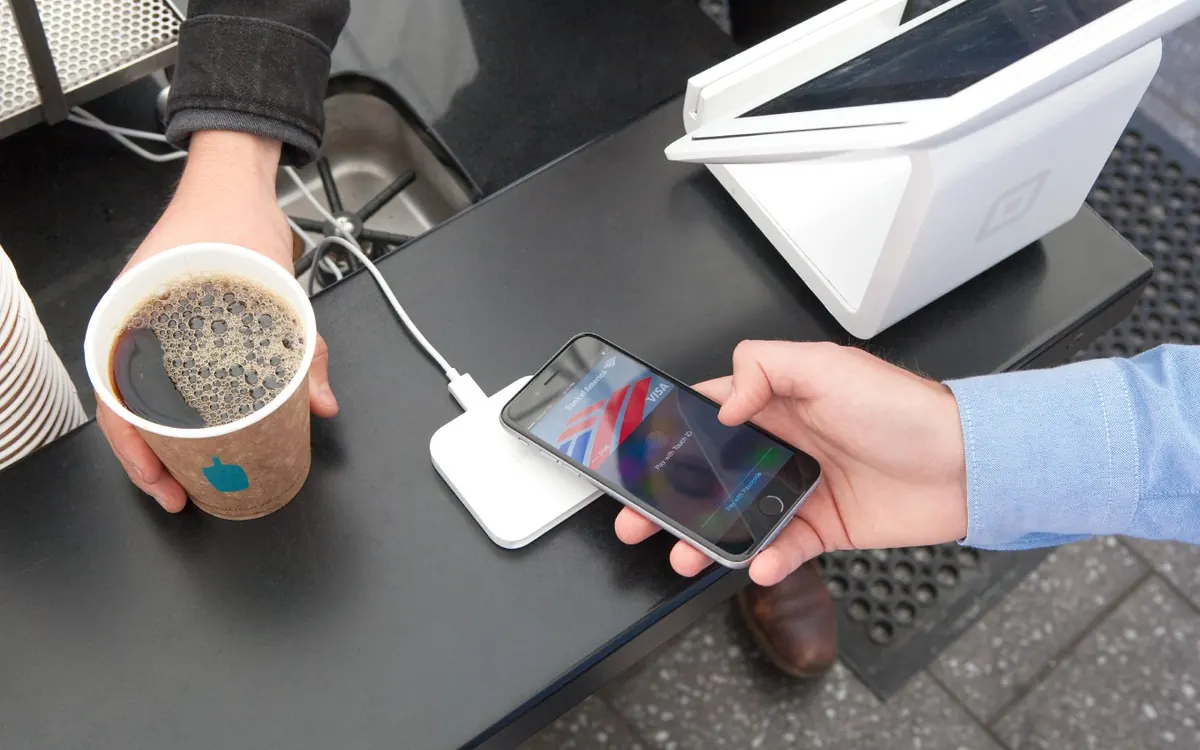 A customer holds an iPhone over a small contactless card reader to pay for a coffee with Apple Pay