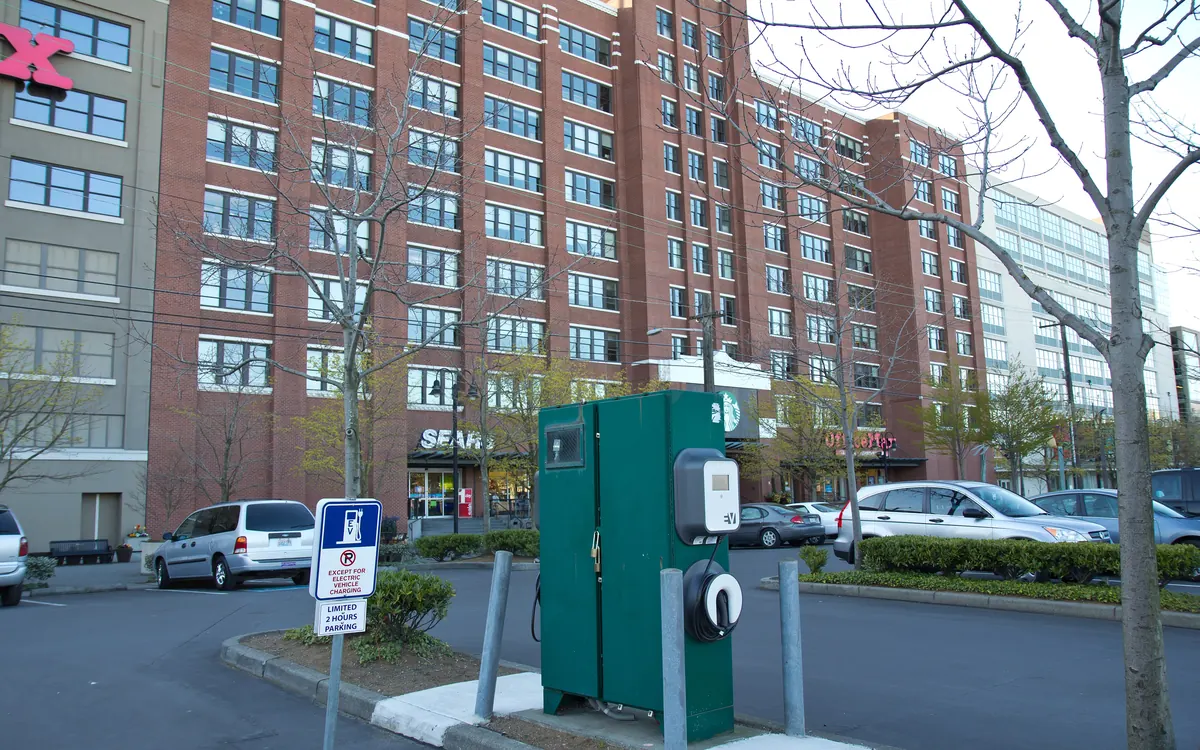 A green public EV charging station beside a parking lot in front of apartment buildings (illustrative photo)