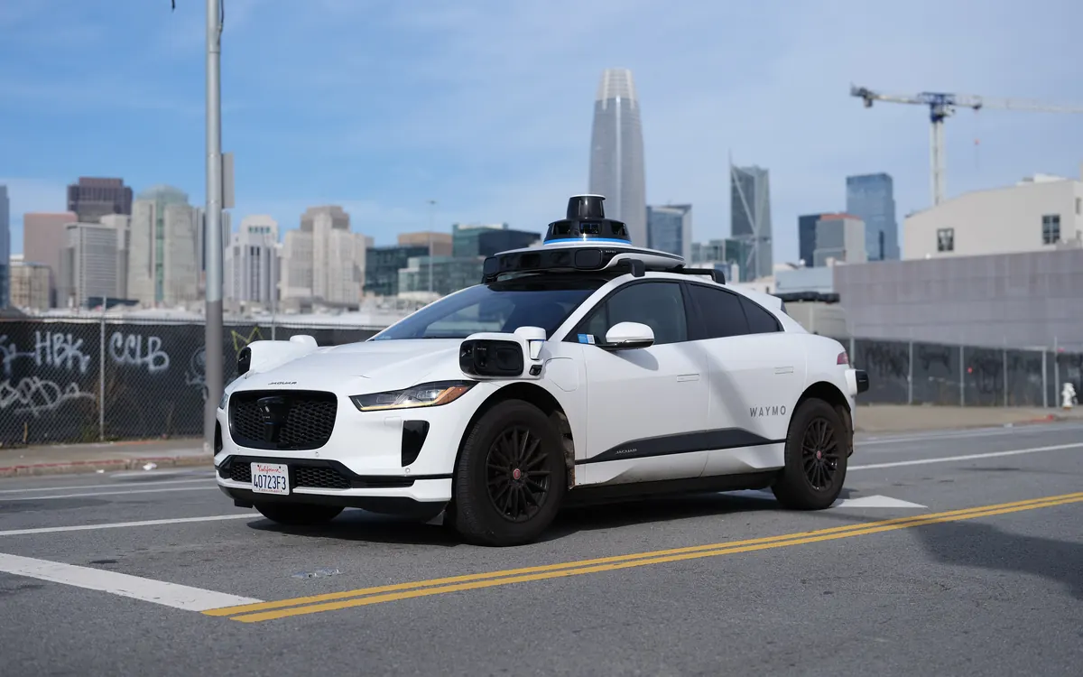 A white Waymo Jaguar I-Pace robotaxi with roof-mounted sensors on a San Francisco street