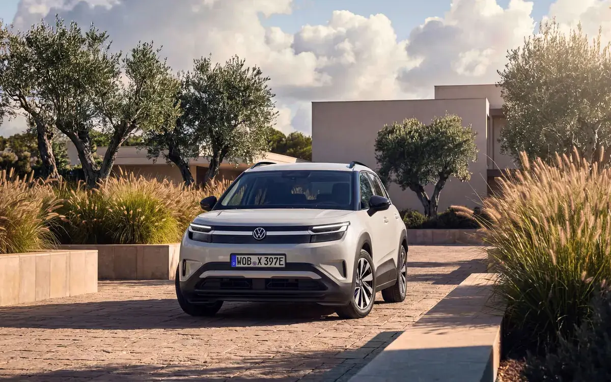 A white Volkswagen ID. Cross compact electric SUV parked on a paved courtyard between olive trees and ornamental grasses